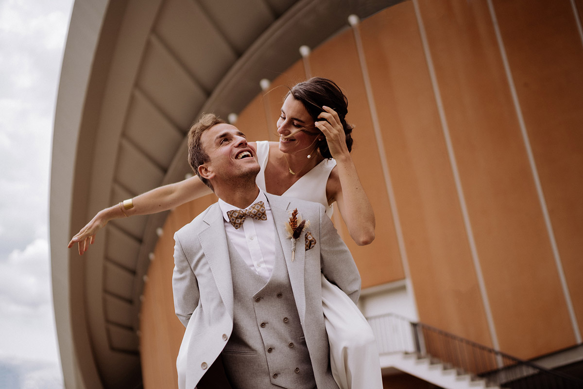 Lachendes Brautpaar beim Huckepack-Shooting vor dem Haus der Kulturen der Welt. Alle Leistungen und Preise der Hochzeitsfotografin Berlin im Überblick.