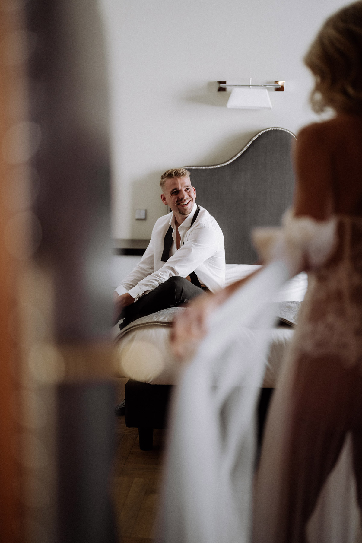 Bräutigam schaut überrascht lachend zur Braut und sitzt auf dem Bett im Hotelzimmer, Getting Ready und Boudoir Stimmung. Foto: Melanie Meißner