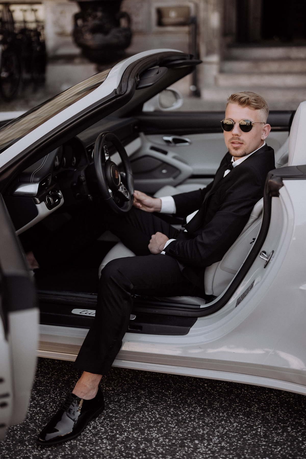 Modernes Fashion-inspiriertes Männerportrait vom Bräutigam mit Sonnenbrille im Hochzeitsauto sitzend