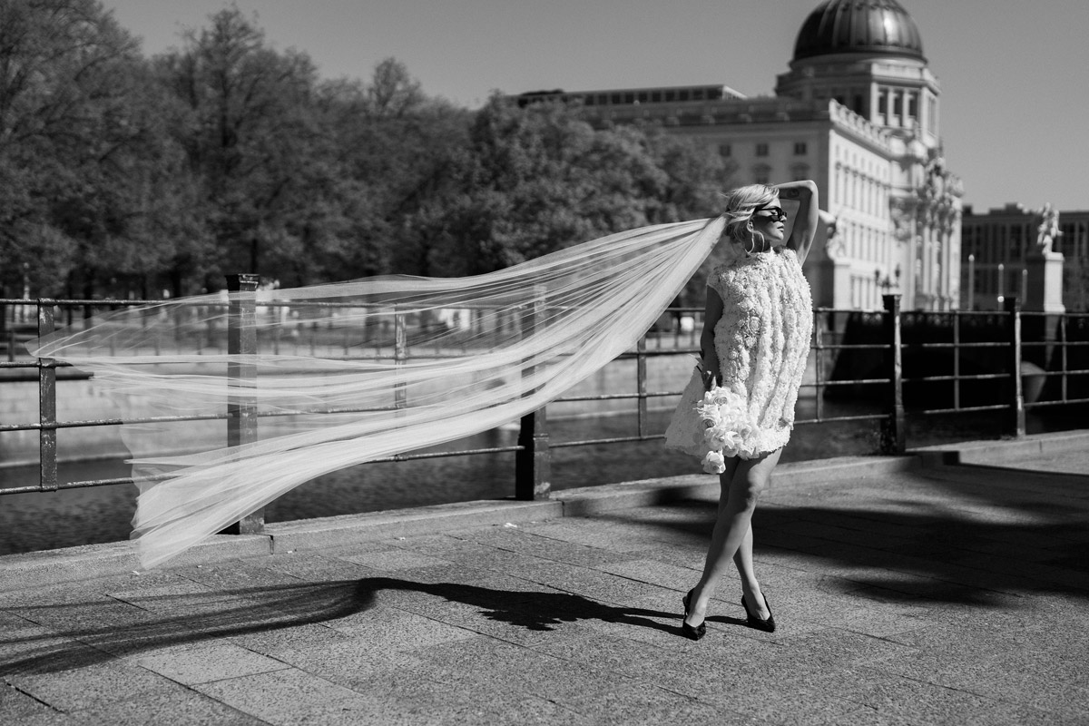 Braut steht mit langem wehendem Schleier an der Spree mit Berliner Schloss im Hintergrund