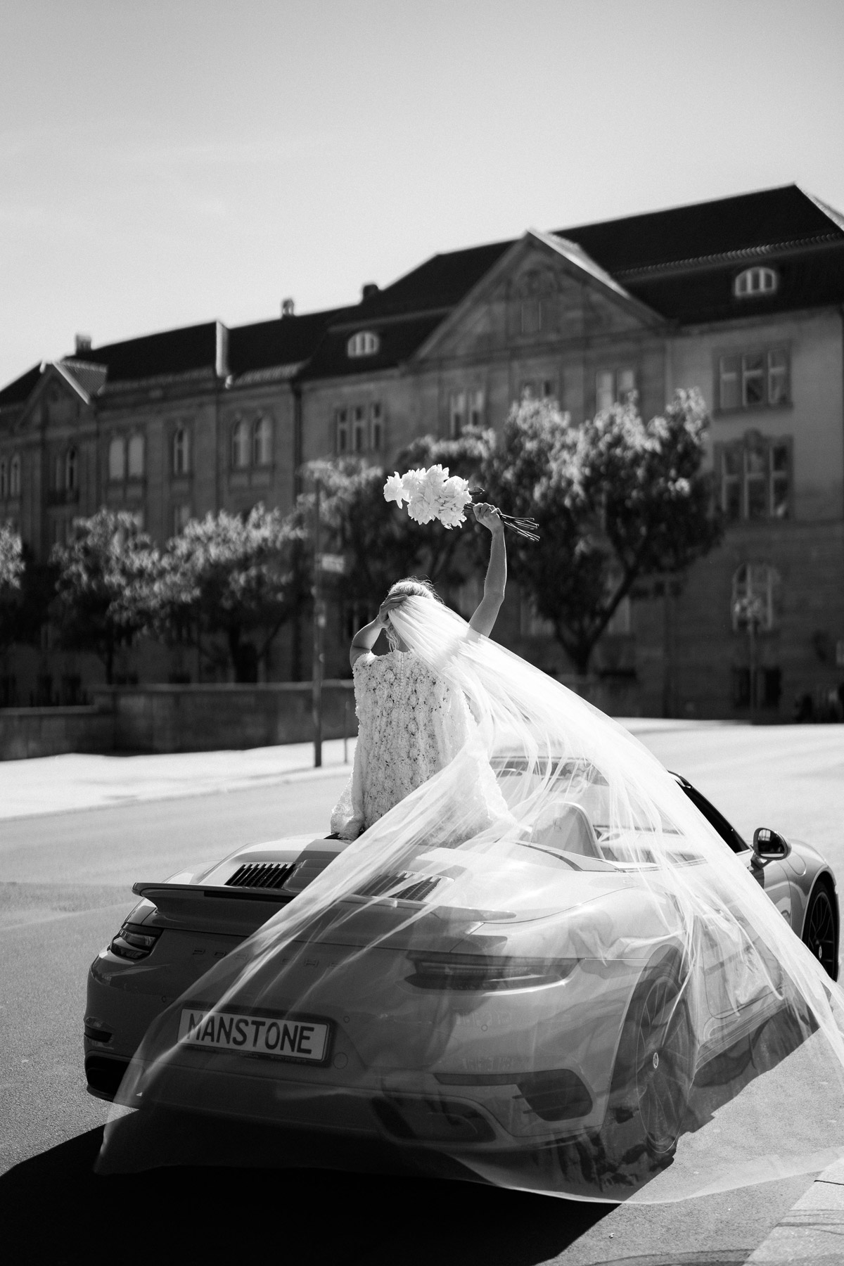 Braut von hinten mit langen, wehendem Schleier sitzt hinten, oben auf Cabrio und wedelt mit Brautstrauß