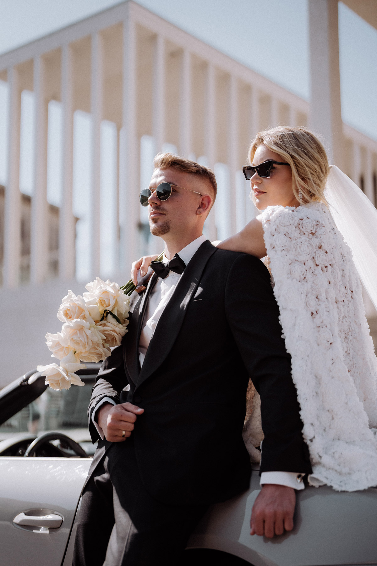 Hochzeitsfotograf Berlin Mitte: Cooles Brautpaar mit Sonnenbrillen steht am Porsche Cabrio vor James-Simon-Galerie