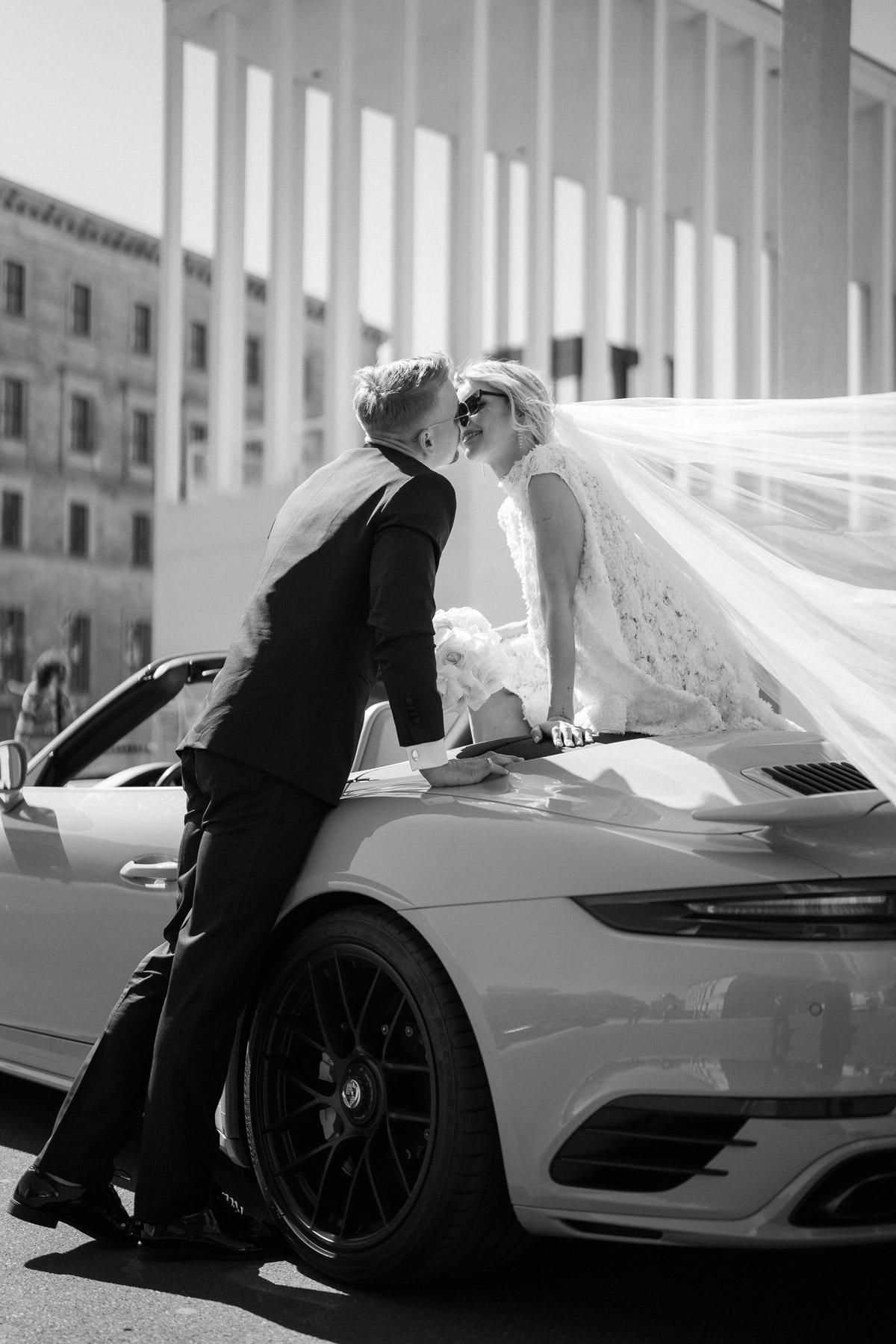 Braut sitzt oben auf Cabrio und küsst Bräutigam