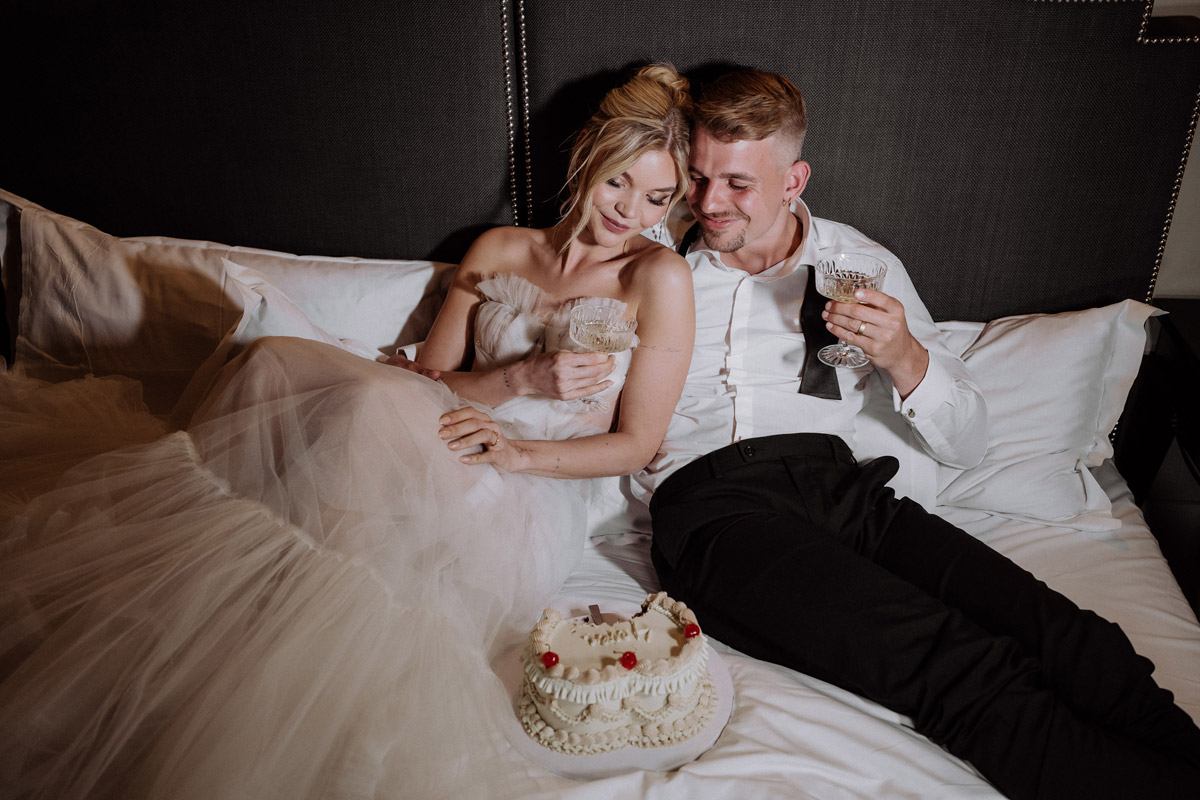 Hochzeitstorte steht im Bett vor dem Elopement-Paar, das Champagner trinkt