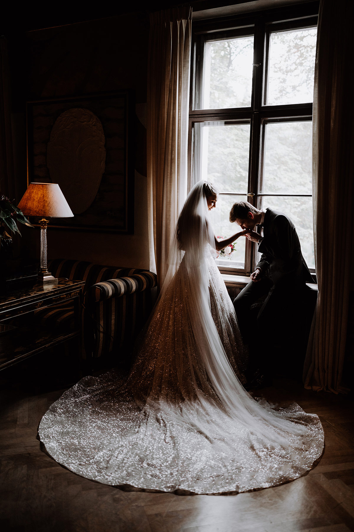 Fotoidee bei Regen im Schlosshotel Berlin: Brautpaar steht am Fenster und Bräutigam küsst Hand der Braut