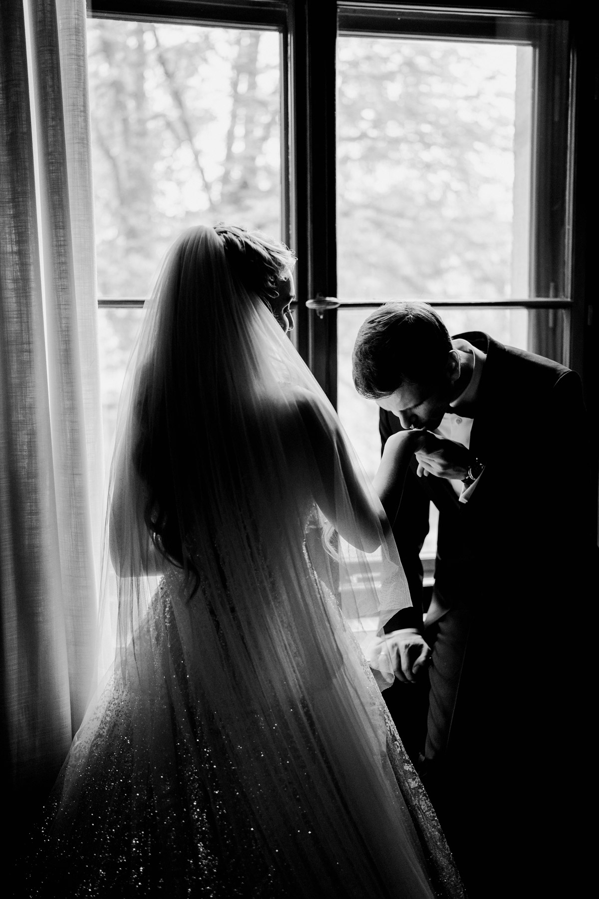 Idee für Hochzeitsfoto im Regen: Hochzeitspaar am Fenster positionieren und Bräutigam gibt Braut Handkuss in Schwarz-Weiß