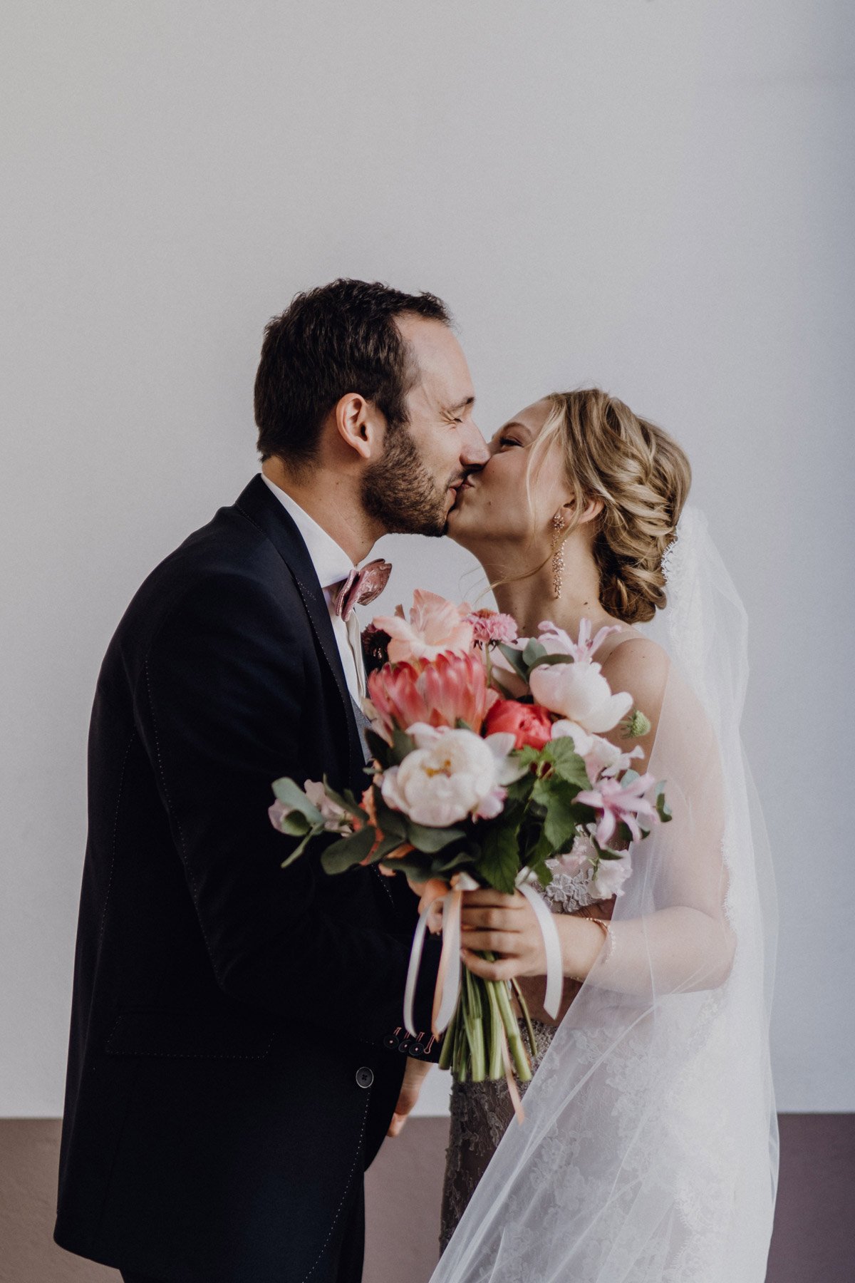 Bräutigam küsst Braut, Fokus liegt auf Boho Brautstrauß in Rosatönen mit Protea von Marsano Blumen