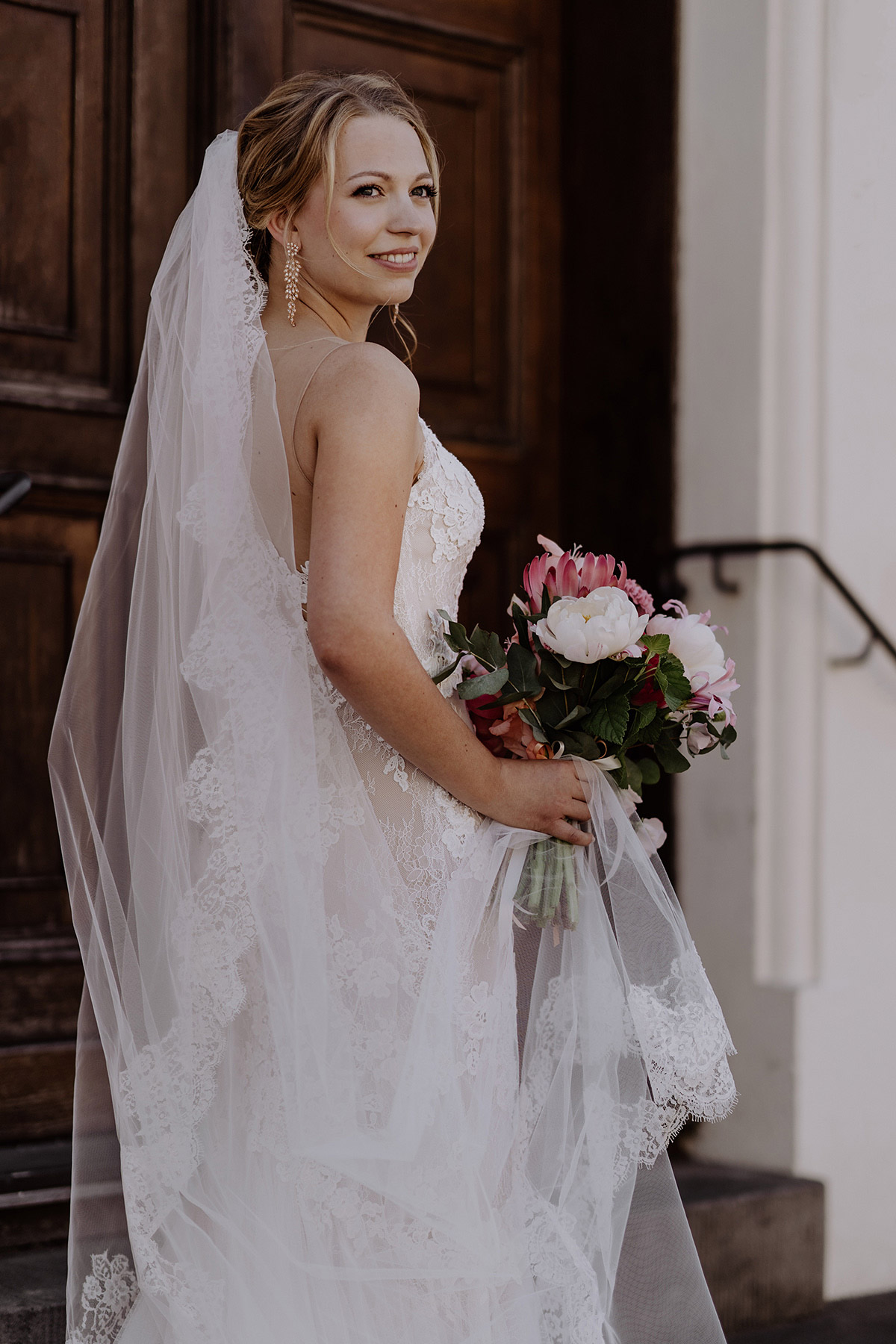 Blonde Braut mit langen Ohrringen und Schleier schaut lächelnd über die Schulter