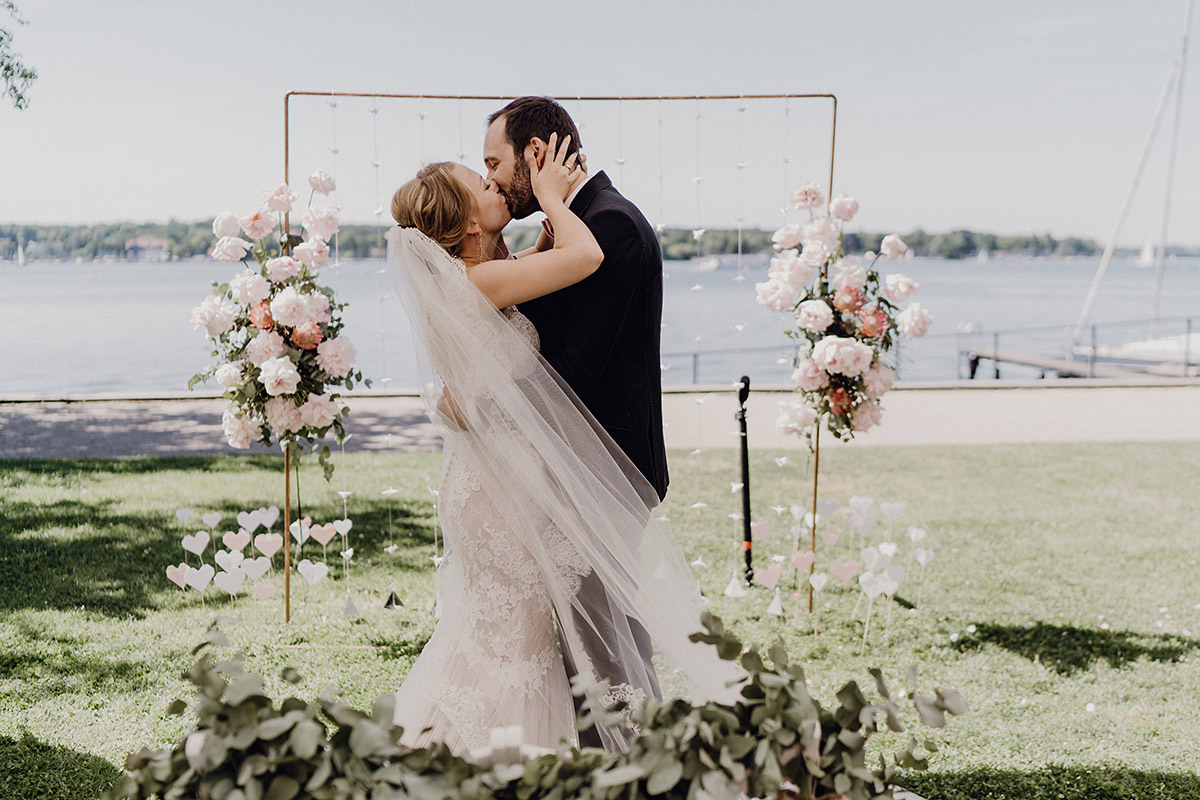 Gästehaus Blumenfisch Hochzeit: Brautpaar küsst sich vor Wannsee-Kulisse und Boho Blumenbogen