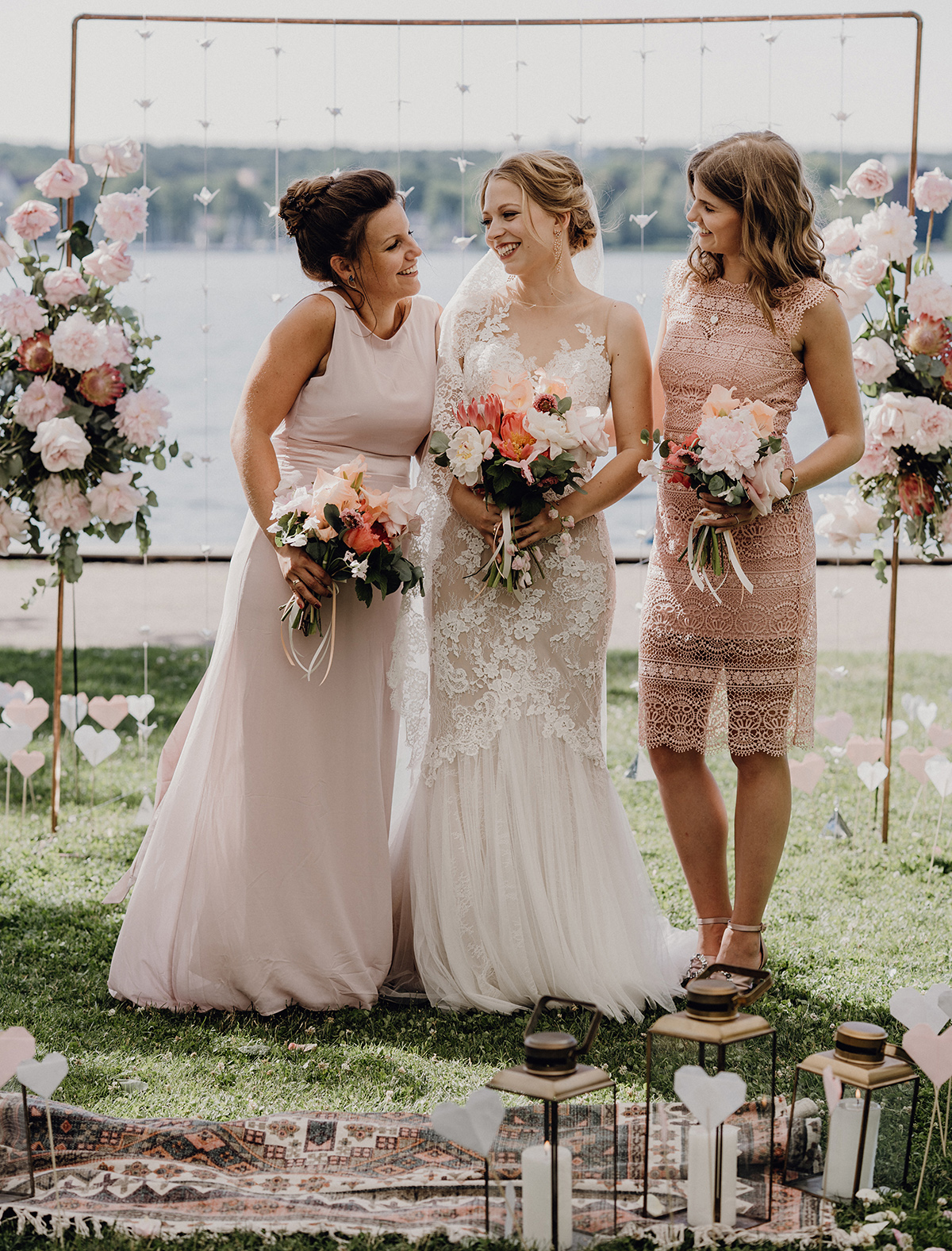 Gruppenfoto: Braut mit Brautjungfern vor Traubogen bei Boho Hochzeit am See