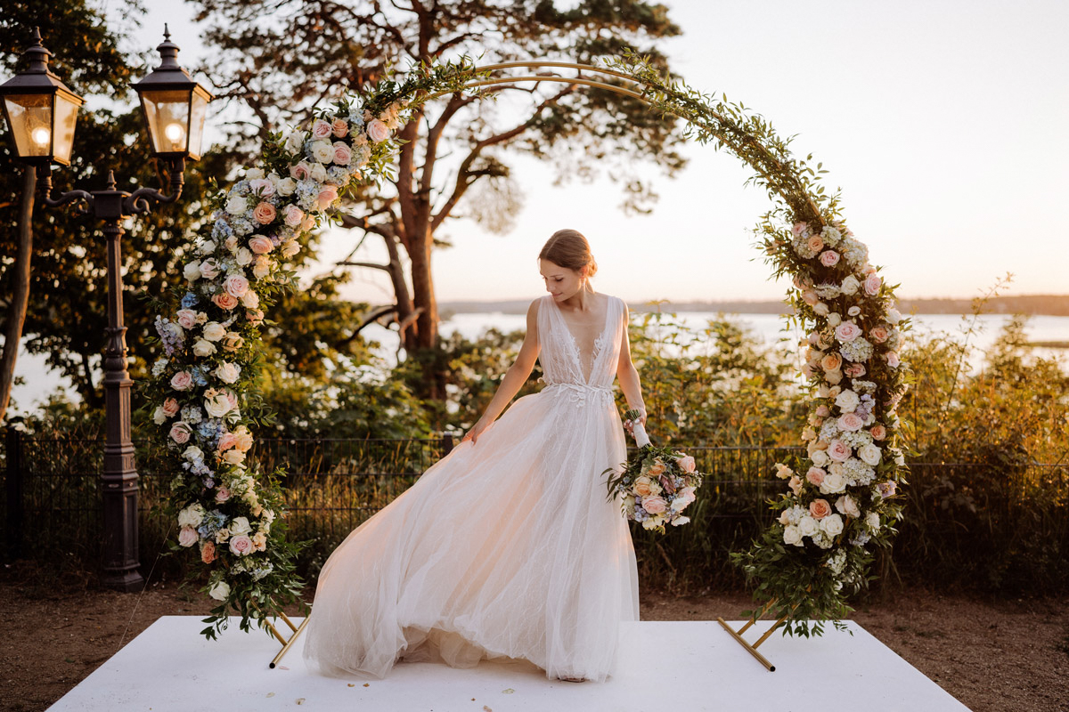 Hochzeitsfotografin Berlin Melanie Meißner (ehemals Hochzeitslicht): Eine elegante Braut steht im goldenen Abendlicht unter einem runden floralen Traubogen an den Wannseeterrassen. Mit über 20 Jahren Erfahrung fange ich solche magischen Momente nun ganz exklusiv für wenige Paare pro Jahr ein.