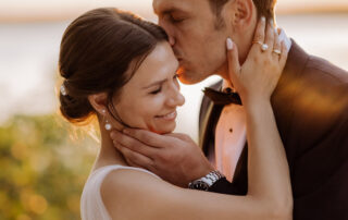 Der Bräutigam küsst die Braut zärtlich auf die Stirn, während sie zur Hochzeit vor der Kulisse des glitzernden Wassers in den Wannseeterrassen Berlin verweilen.