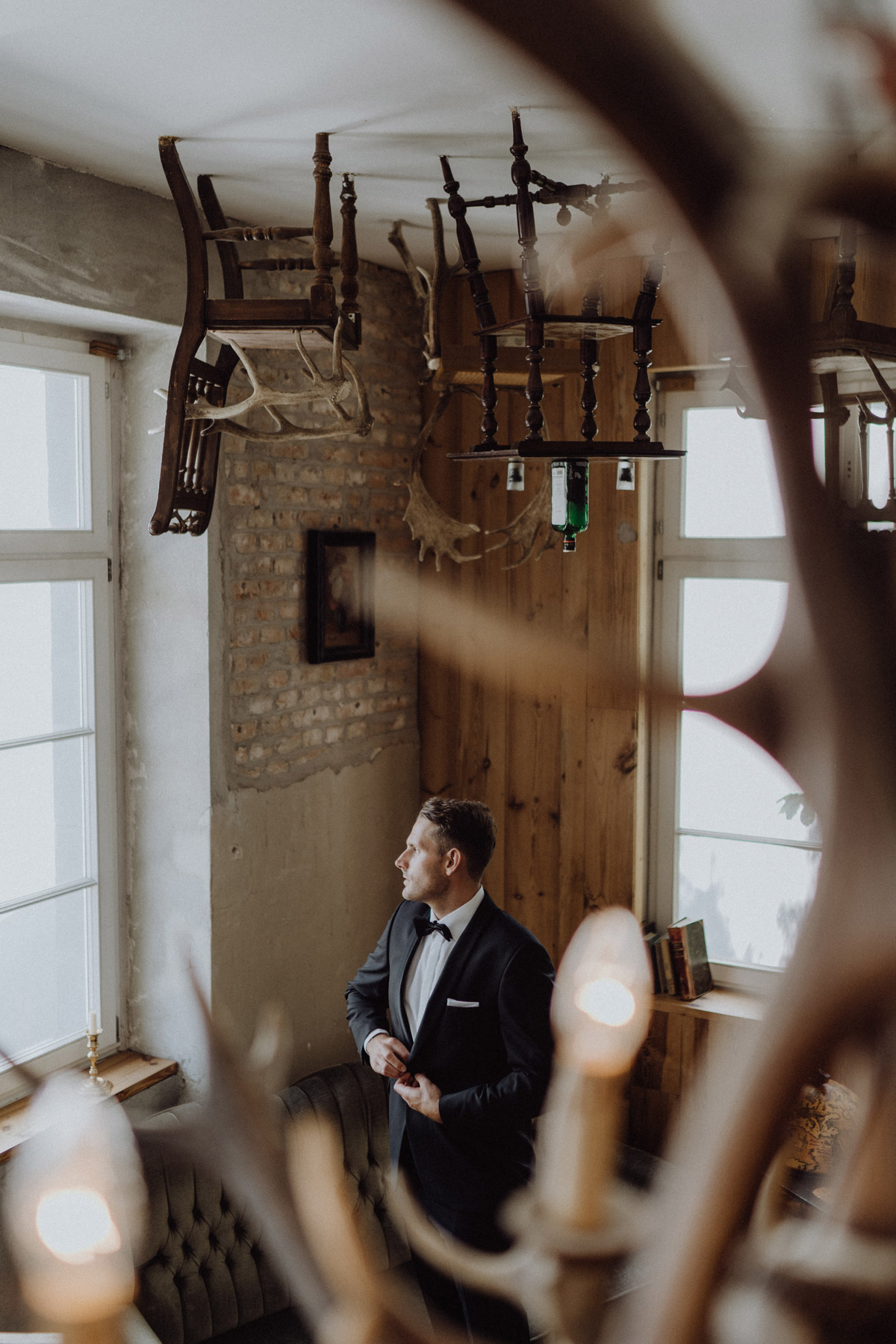 Bräutigam zieht sein Jackett an: Vorbereitungen zur Hochzeit im The Grand Berlin im kreativen Raum mit Geweihen, Tisch und Stühlen an der Decke