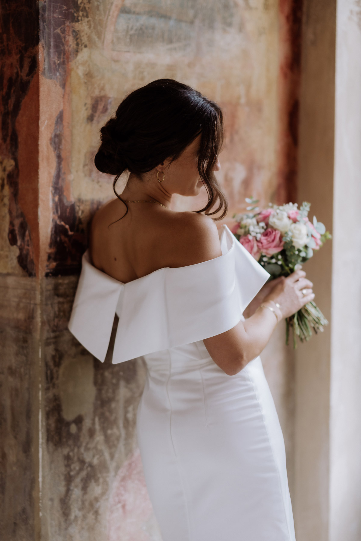 Braut in Vogue Pose: Mit schulterfreiem Kleid und Hochsteckfrisur von hinten steht sie verträumt am Fenster.