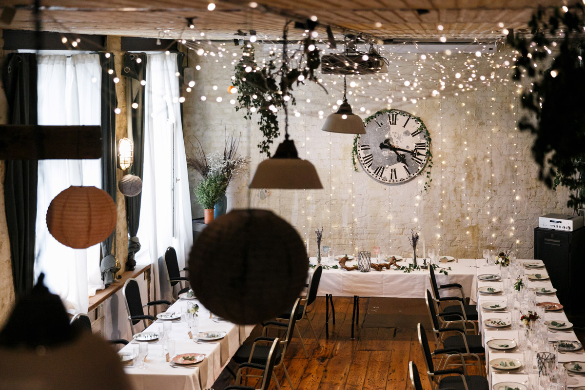 Hochzeitslocation Old Smithy's Dizzle Berlin: Stimmungsvoll beleuchteter Feierraum mit Lichterketten, Vintage-Uhr und festlich gedeckten Tafeln im Industrie-Chic