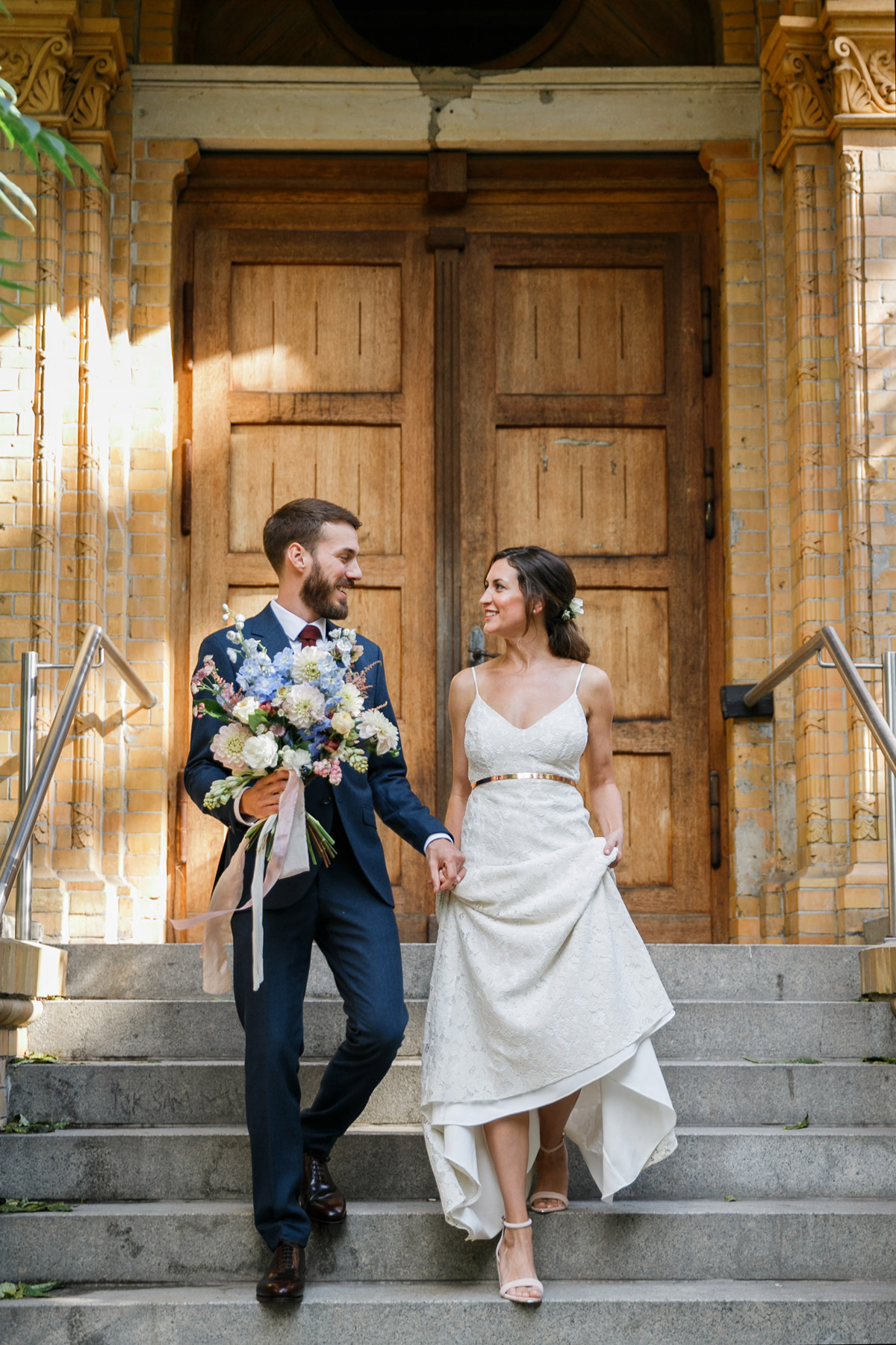 Entspanntes Brautpaarshooting in Berlin: Braut und Bräutigam laufen lachend und Hand in Hand die Steintreppe vor einer historischen Holztür hinab