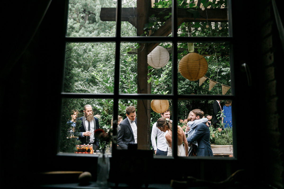 Hochzeitsreportage Berlin Friedrichshain: Blick durch ein historisches Sprossenfenster auf die entspannte Hochzeitsgesellschaft beim Sektempfang im Garten
