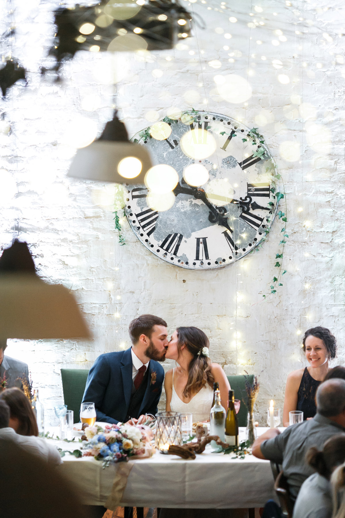 Hochzeitsfeier Berlin Friedrichshain: Romantischer Kuss des Brautpaares am Hochzeitstisch unter der großen Vintage-Uhr im Old Smithy's Dizzle