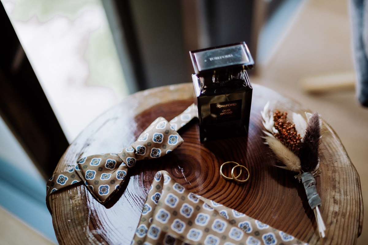 Flatlay mit Details: Parfüm, Fliege, Eheringe, Einstecktuch und Ansteckstrauß des Bräutigams