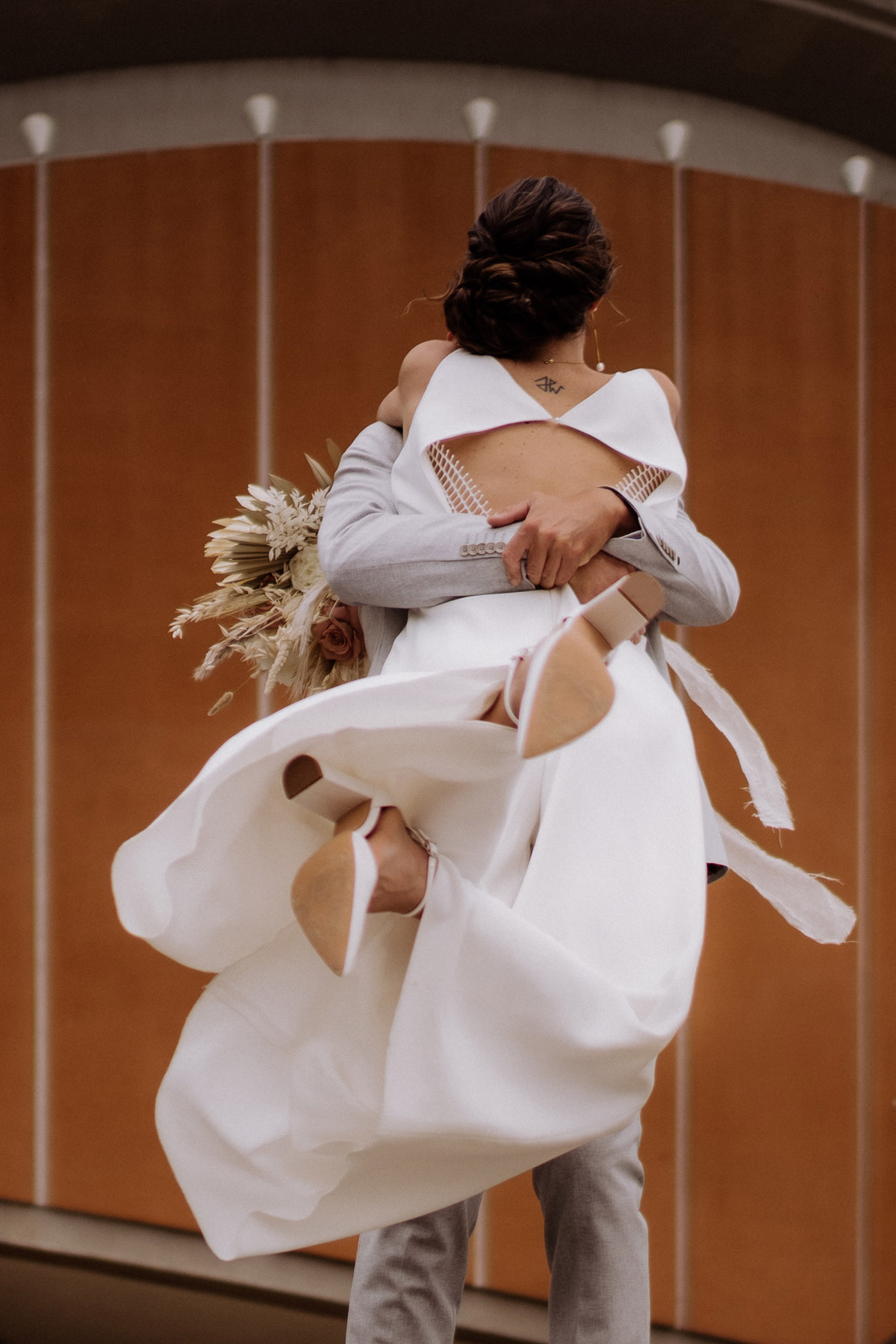 Emotionaler Moment beim Hochzeitsshooting: Bräutigam hebt Braut hoch, Detailansicht des Jumpsuits vor der „Schwangeren Auster“