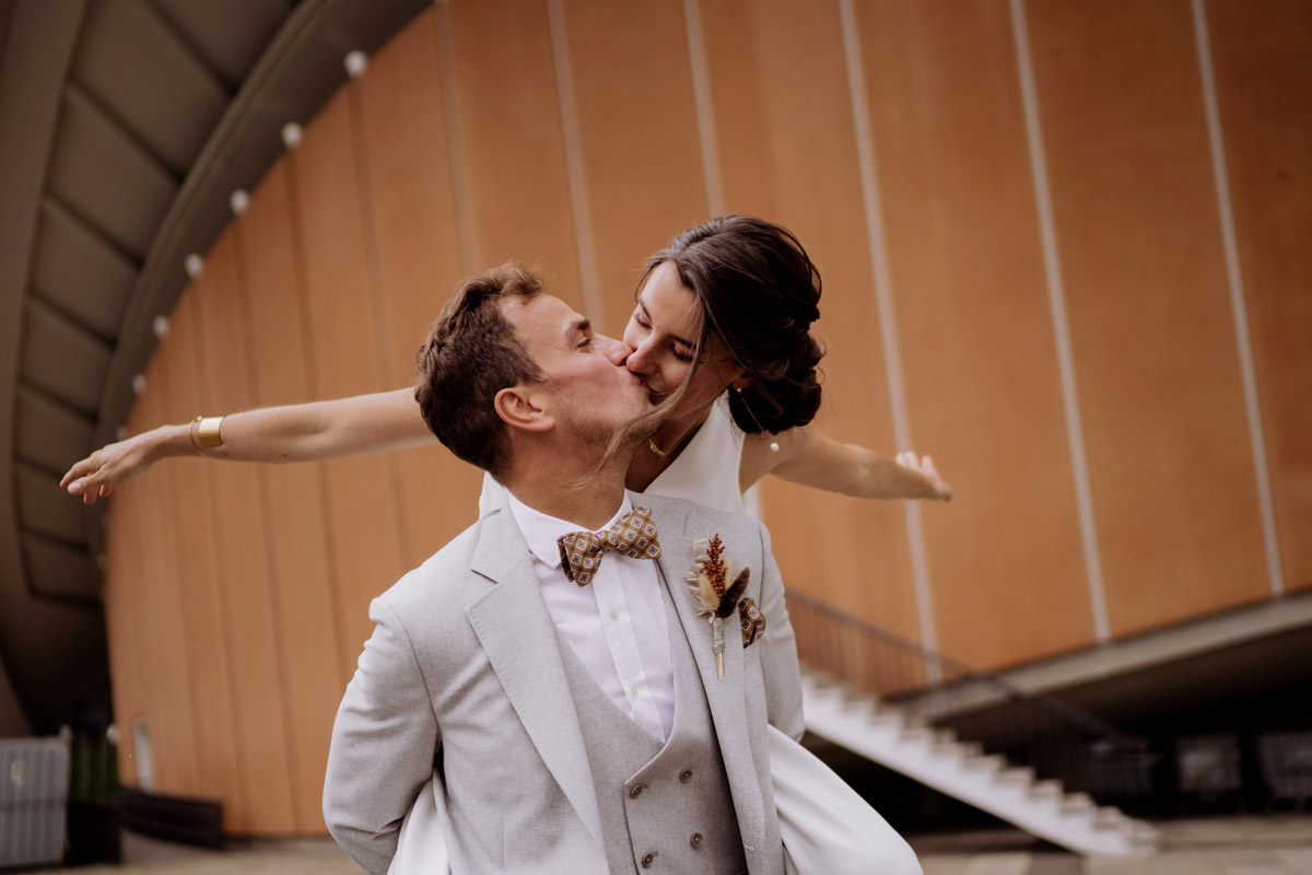 Kreatives Hochzeitsfotoshooting: Braut auf dem Rücken des Bräutigams streckt Arme in Fliegerposition aus und beide küssen sich