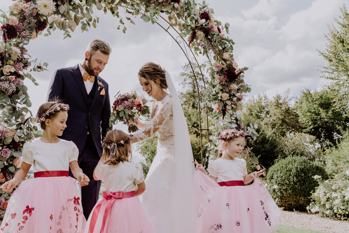 Hochzeitsfotograf Potsdam: Glückliches Brautpaar und kleine Blumenmädchen am prachtvollen Traubogen bei der freien Trauung auf Gut Schloss Golm