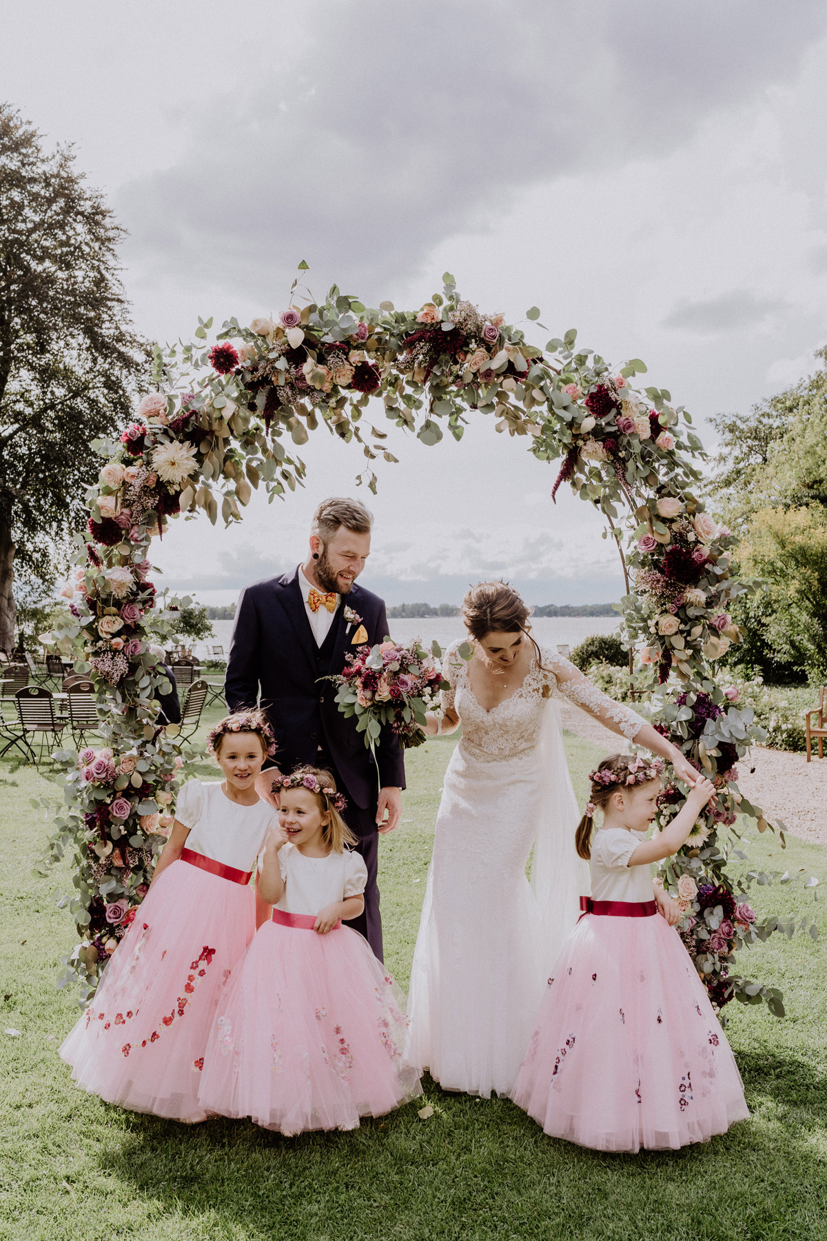 Hochzeitsfotograf Potsdam: Lachendes Brautpaar mit Blumenmädchen vor der traumhaften Seekulisse auf Gut Schloss Golm