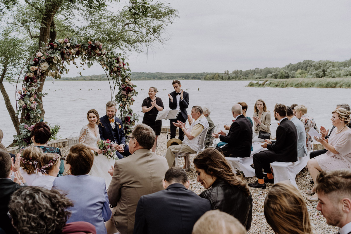 Hochzeitslocation Brandenburg am Wasser: Hochzeitsgesellschaft klatscht für das Brautpaar bei der idyllischen Schlosshochzeit direkt am Seeufer