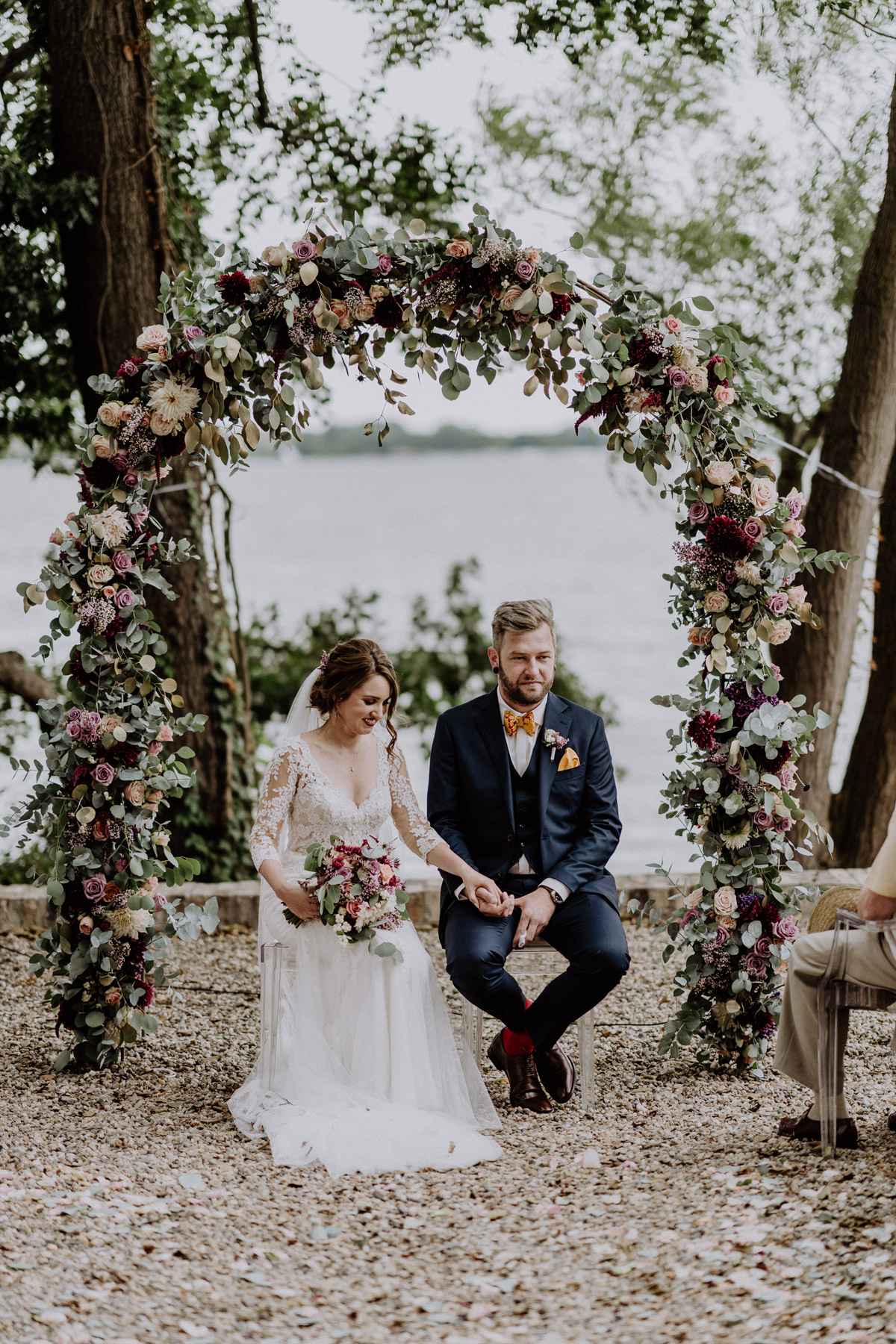 Heiraten am Wasser in Potsdam: Brautpaar sitzt strahlend unter dem üppigen Blumenbogen während der emotionalen Hochzeitszeremonie am See