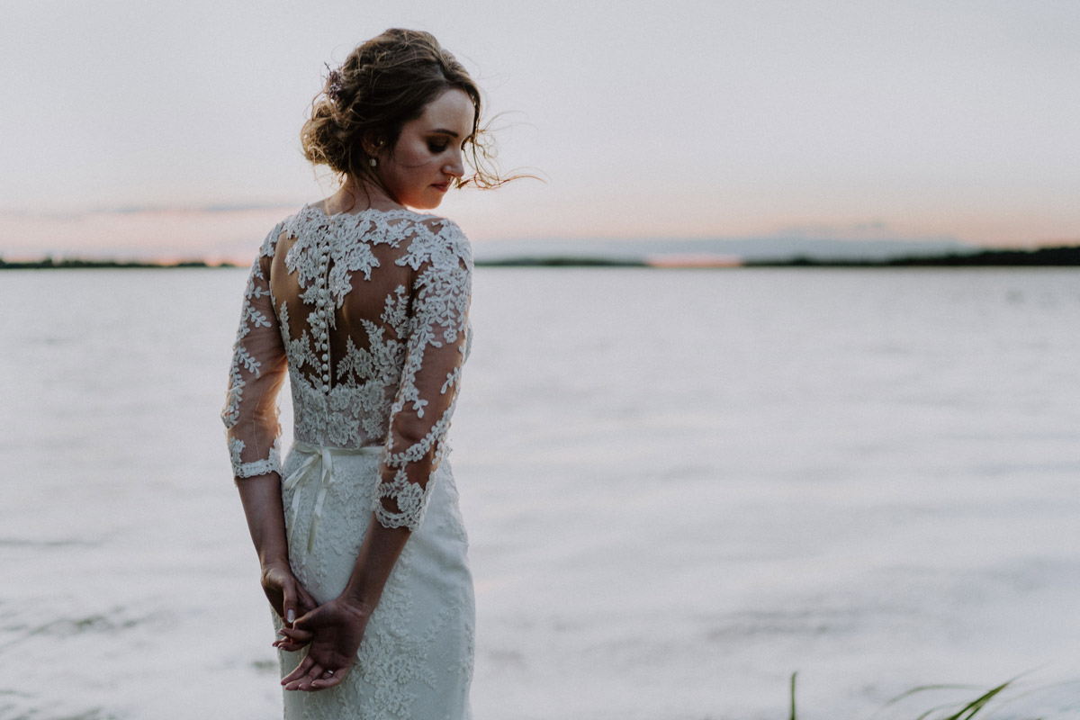 Elegante Braut im rückenfreien Spitzenkleid genießt den romantischen Sonnenuntergang am See