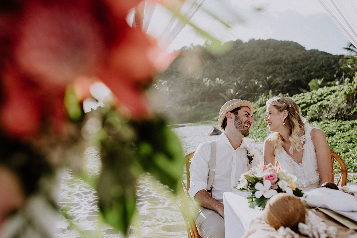 Destination Wedding: Das Brautpaar aus Berlin lacht ausgelassen während der intimen Hochzeitszeremonie am tropischen Strand, fotografiert durch exotische Blumen