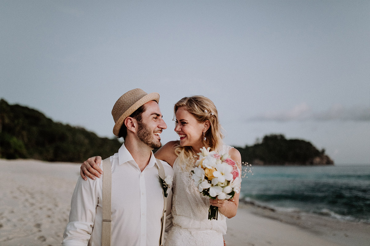 Hochzeitsfotograf Charlottenburg goes Seychellen: Die Braut mit tropischem Brautstrauß und ihr Bräutigam blicken sich beim Elopement-Shooting am weißen Strand verliebt an