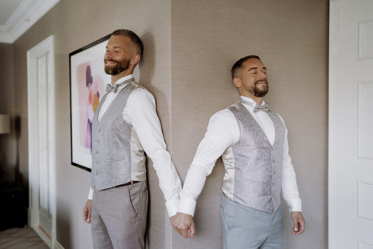 emotionaler First Look Moment der schwulen Hochzeit: beide Bräutigame halten sich an der Hand und können sich noch nicht sehen