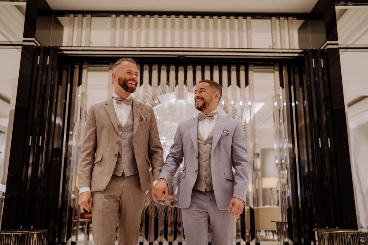Männer-Hochzeit: Bräutigame laufen Hand in Hand und freudestrahlend durch das Hotel