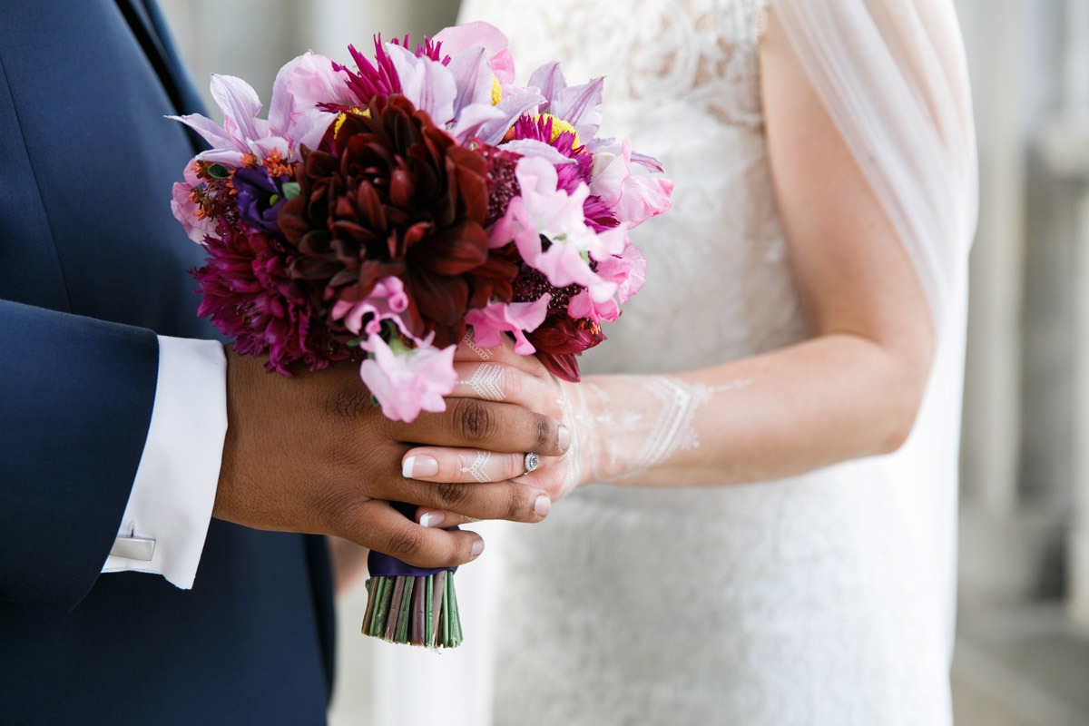Detailaufnahme einer internationalen Hochzeit: Brautpaar hält sich an den Händen, Fokus auf Brautstrauß in Beerentönen und weißem Henna-Tattoo der Braut