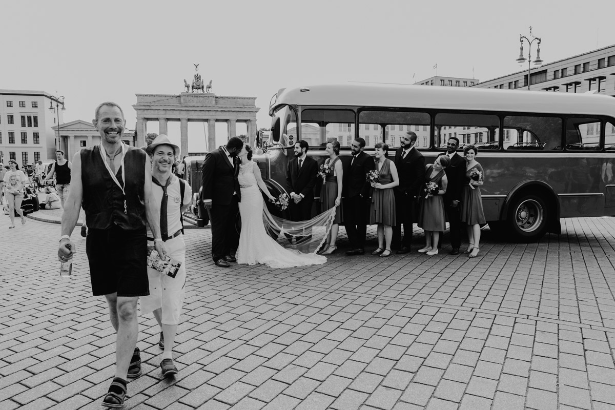 Hochzeitsreportage in Schwarz-Weiß: lachende Touristen laufen im Gruppenfoto mit Oldtimer Bus vor Brandenburger Tor