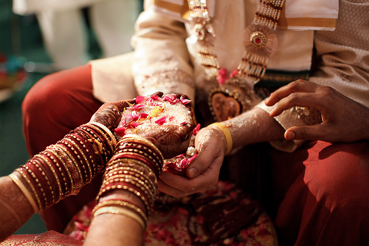 Hindu Wedding Photographer Berlin: Close-up der Hände von Braut und Bräutigam, die Reis und rote Blütenblätter für das traditionelle Hochzeitsritual halten. Braut mit kunstvollem Henna Mehndi und indischen Armreifen