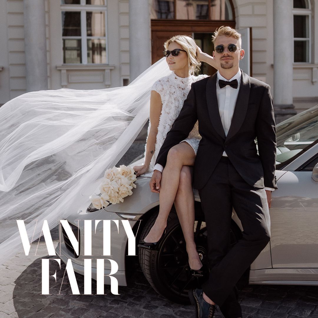 Stilvolles Brautpaar mit Sonnenbrillen posiert lässig auf einem Sportwagen, der Schleier der Braut weht im Wind. Hochzeitsfotografie im Fashion- und Editorial-Look mit Vanity Fair Schriftzug.