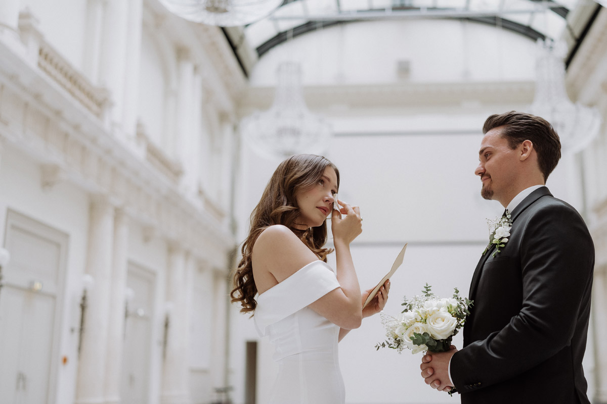 Emotionale Hochzeitsreportage: Die Braut wischt sich beim Vorlesen des Ehegelübdes eine Träne aus dem Gesicht, während der Bräutigam sie liebevoll anlächelt. Authentische Momentaufnahme einer Hochzeitsfotografin in der eleganten Location Hotel de Rome Festsaal in Berlin.
