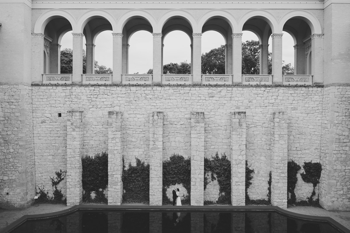 Künstlerische Weitwinkelaufnahme im Innenhof des Belvedere Pfingstberg in Potsdam: Das Brautpaar ist klein in der monumentalen Architektur zu sehen. Ein Beispiel für die cineastische Bildkomposition und den individuellen Schnitt eures Hochzeitsfilms.