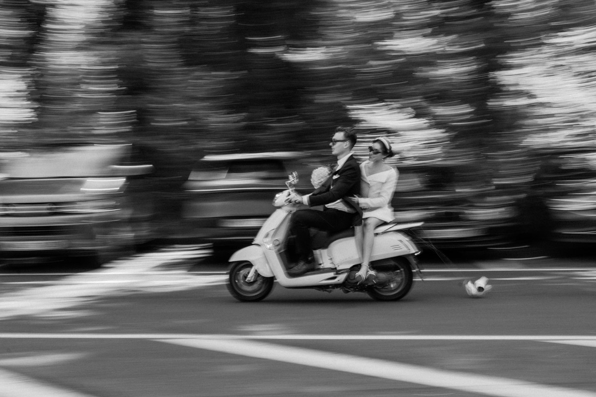 Mitzieher Hochzeitsfoto in Schwarz-Weiß. Das Paar fährt auf einer Vespa zu seiner Rathaus Standesamt Tempelhof-Schöneberg Hochzeit. FAQ zur Mindestbuchungsdauer für Hochzeitsfotografie Berlin.