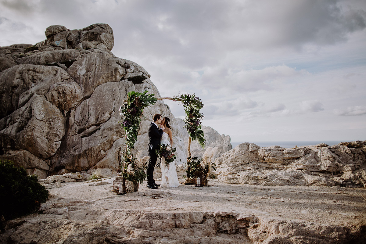 Emotionale Hochzeitsfotografie auf Mallorca: Das Brautpaar küsst sich während der freien Trauung unter einem floralen Traubogen. Ein Beispiel für die vollständige Bildauswahl und Dokumentation wichtiger Momente.