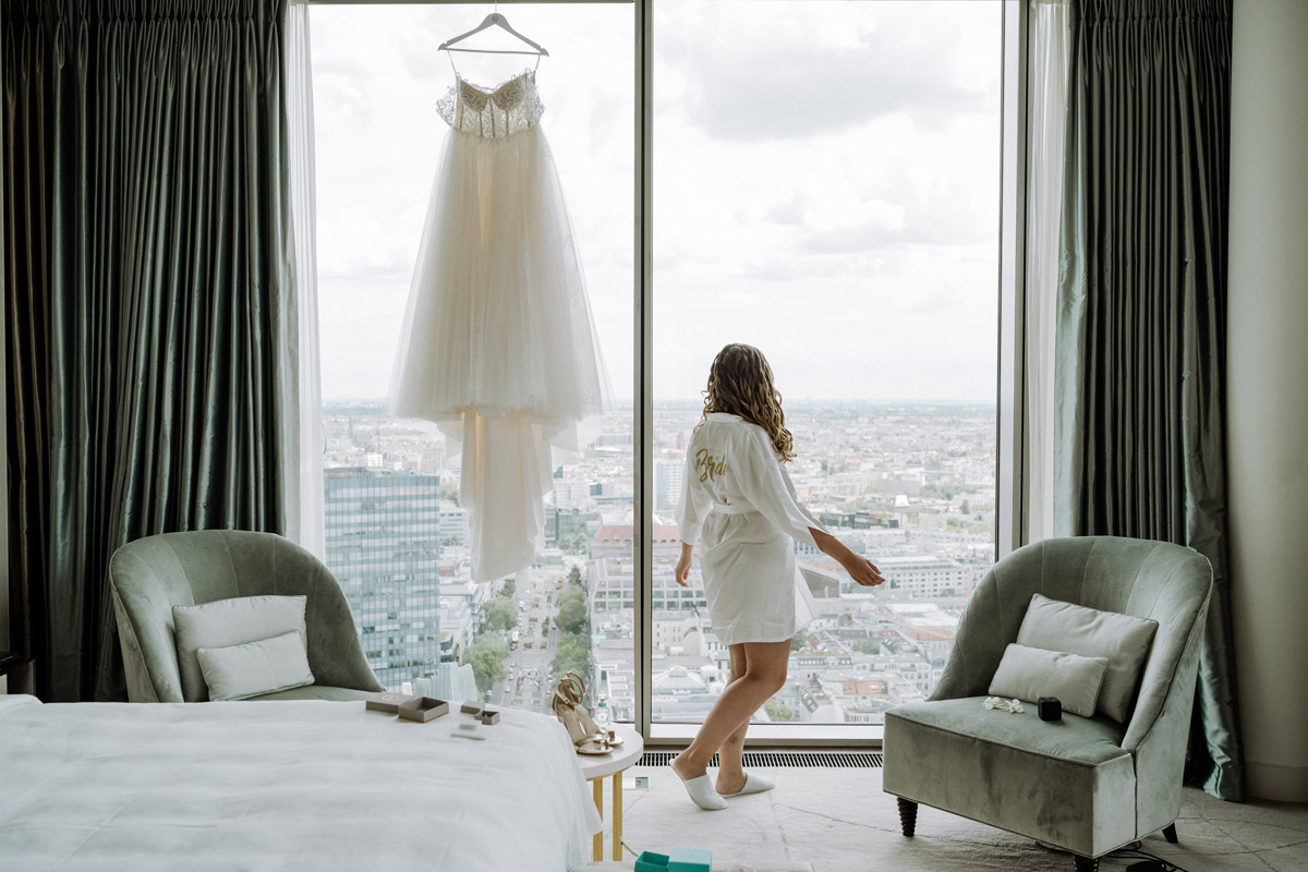 Cineastisches Getting Ready im Waldorf Astoria Berlin: Die Braut im Bademantel blickt auf die Berliner Skyline, während das Hochzeitskleid am Fenster hängt. Ein Ausblick auf die emotionale Story und Länge eures Hochzeitsvideos.