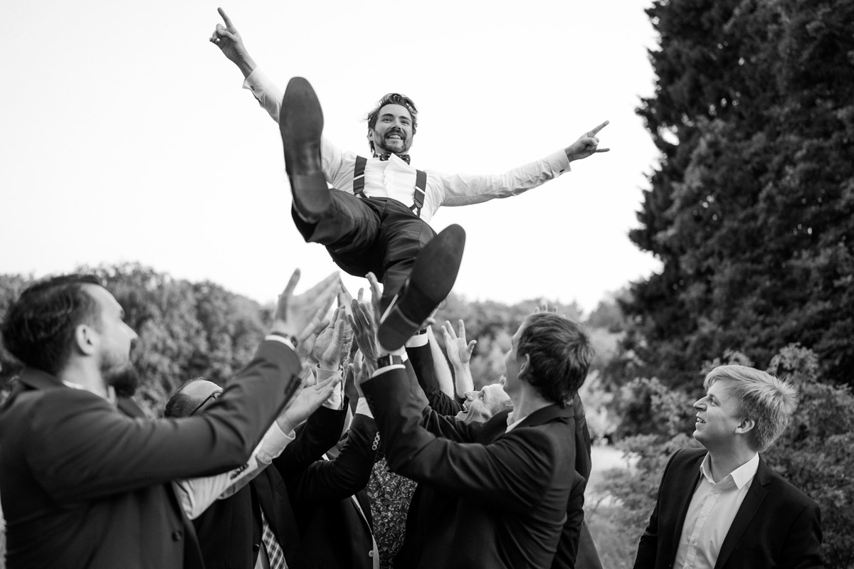 Ausgelassene Stimmung beim Gruppenfoto-Shooting: Der lachende Bräutigam wird von seinen Freunden und Trauzeugen in die Luft geworfen. Ein dynamisches und kreatives Gruppenbild in Schwarz-Weiß von Hochzeitsfotografin Berlin.