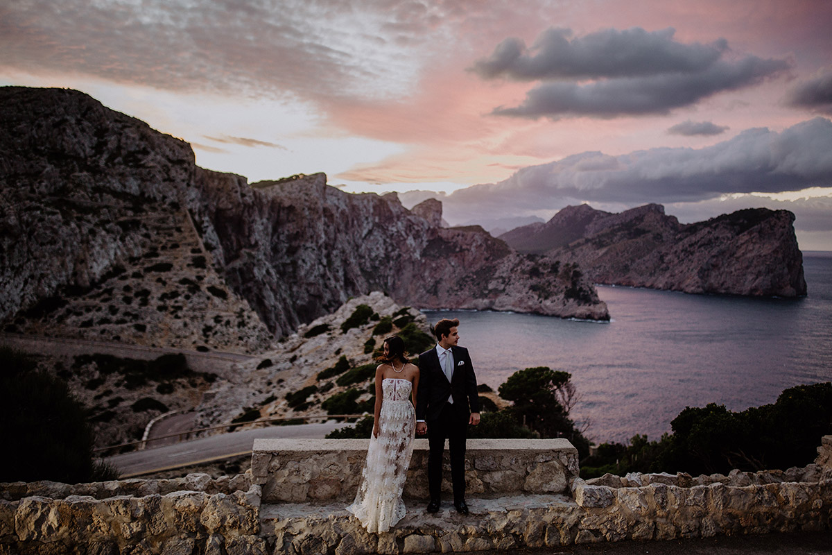 Malerische Hochzeitsfotografie: Das Brautpaar steht bei Sonnenuntergang an einer Steilküste auf Mallorca mit weitem Blick über das Meer. Ein kunstvoller Moment, der die Synergie von Foto und Video bei einer Destination Wedding zeigt.