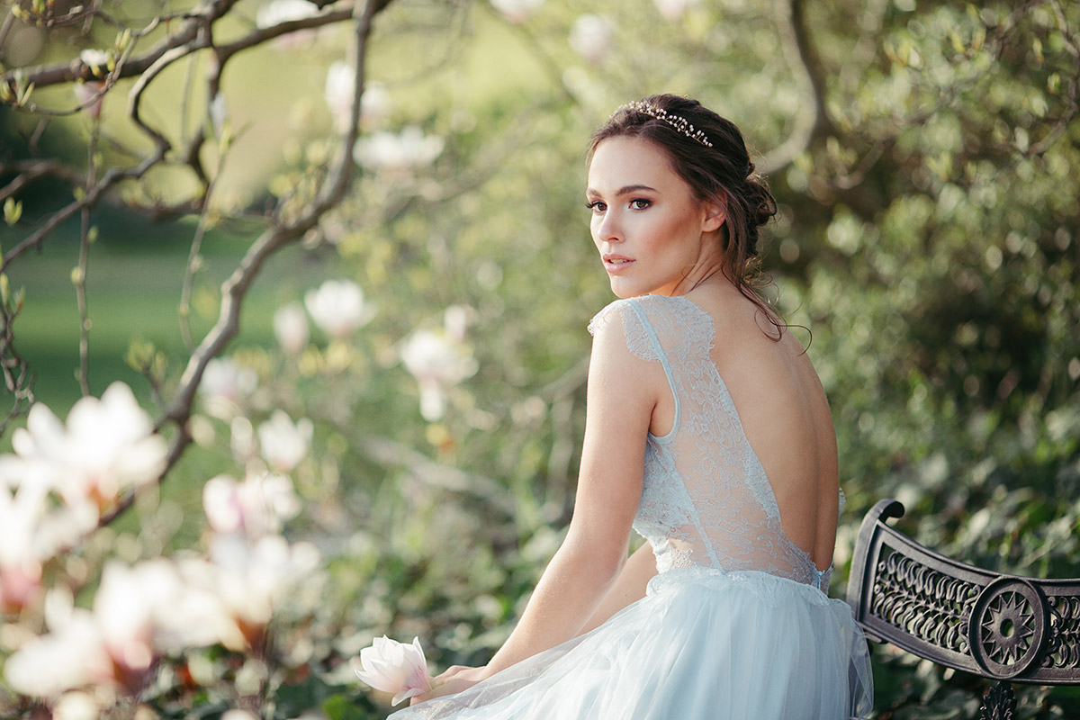 Nachhaltige Brautmode in Berlin: Braut im hellblauen Second-Hand-Kleid sitzt unter blühenden Magnolien auf einer Holzbank. Authentische Hochzeitsfotografie, die ressourcenschonende Ansätze und Natürlichkeit betont.