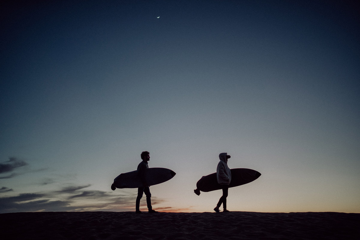 Künstlerisches Silhouetten-Foto eines Paares, das getrennt hintereinander mit Surfbrettern im Mondlicht am Strand spaziert. Informationen zu Stornierung und Verschiebung einer Hochzeit bei deiner Hochzeitsfotografin Berlin.