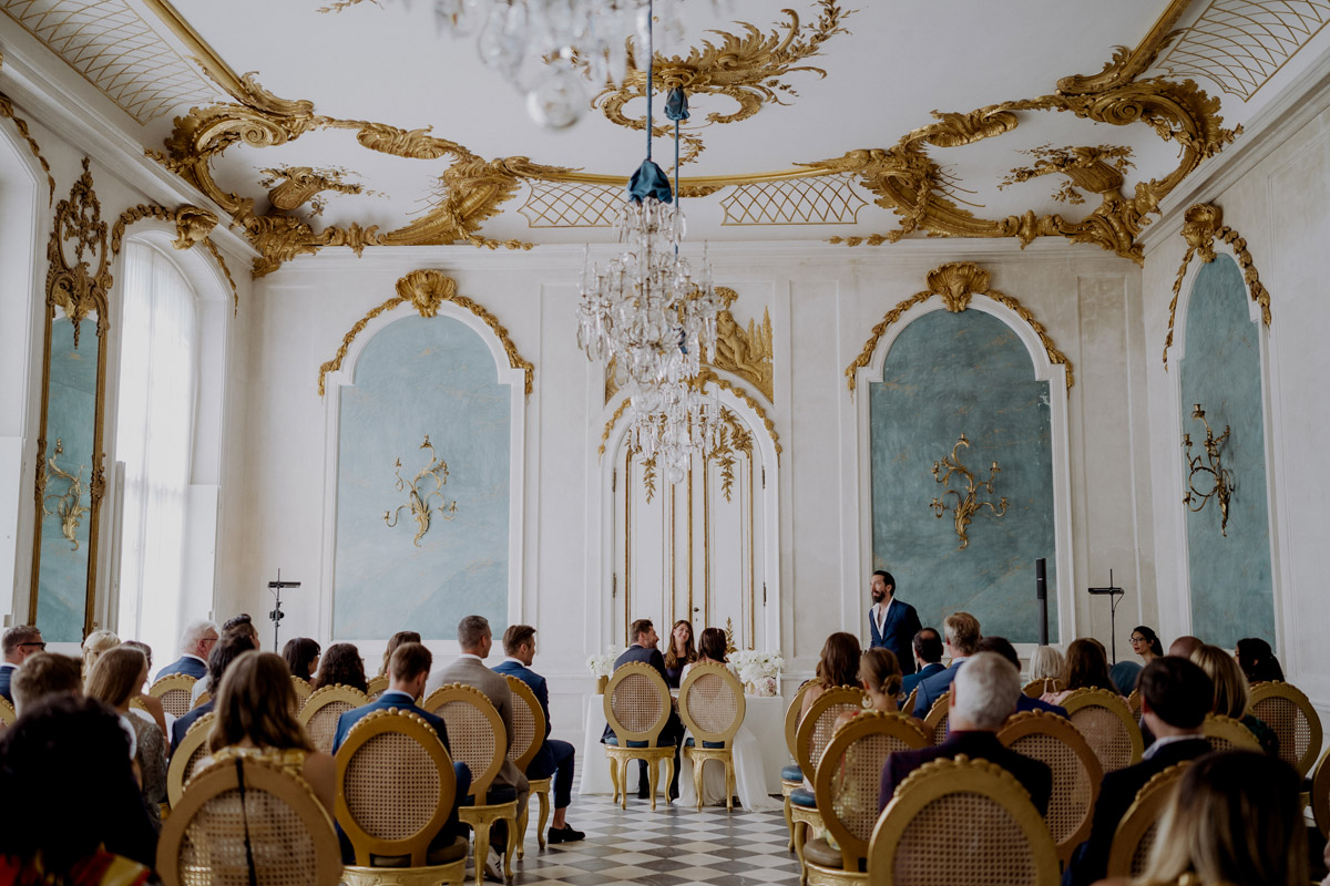 Elegante standesamtliche Trauung im prunkvollen Saal der Neuen Kammern von Sanssouci in Potsdam. Das Brautpaar sitzt während der Zeremonie in der Blauen Galerie vor seinen Hochzeitsgästen.