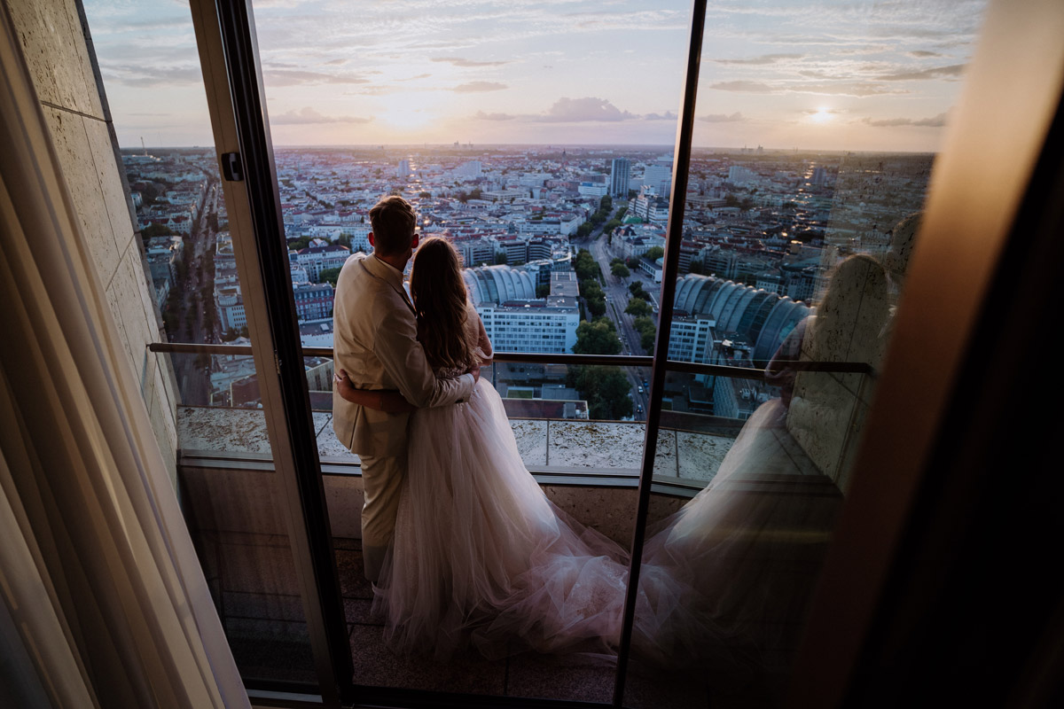Romantisches Brautpaar am Panoramafenster mit Blick über die Skyline von Berlin bei Sonnenuntergang. Stimmungsvolle Hochzeitsfotografie während einer Rooftop-Hochzeit im Waldorf Astoria.